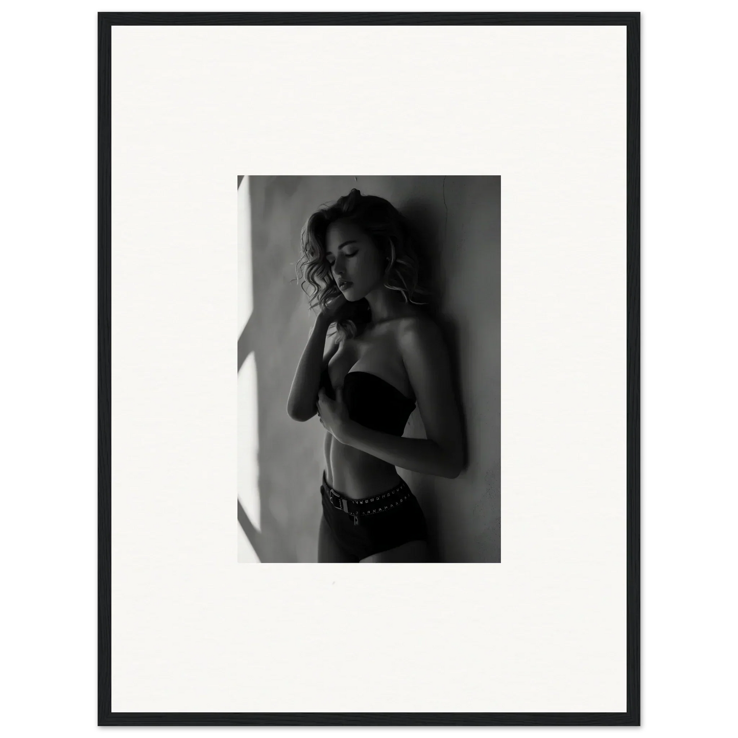 Black and white photograph of a woman in a strapless top posing against a wall.