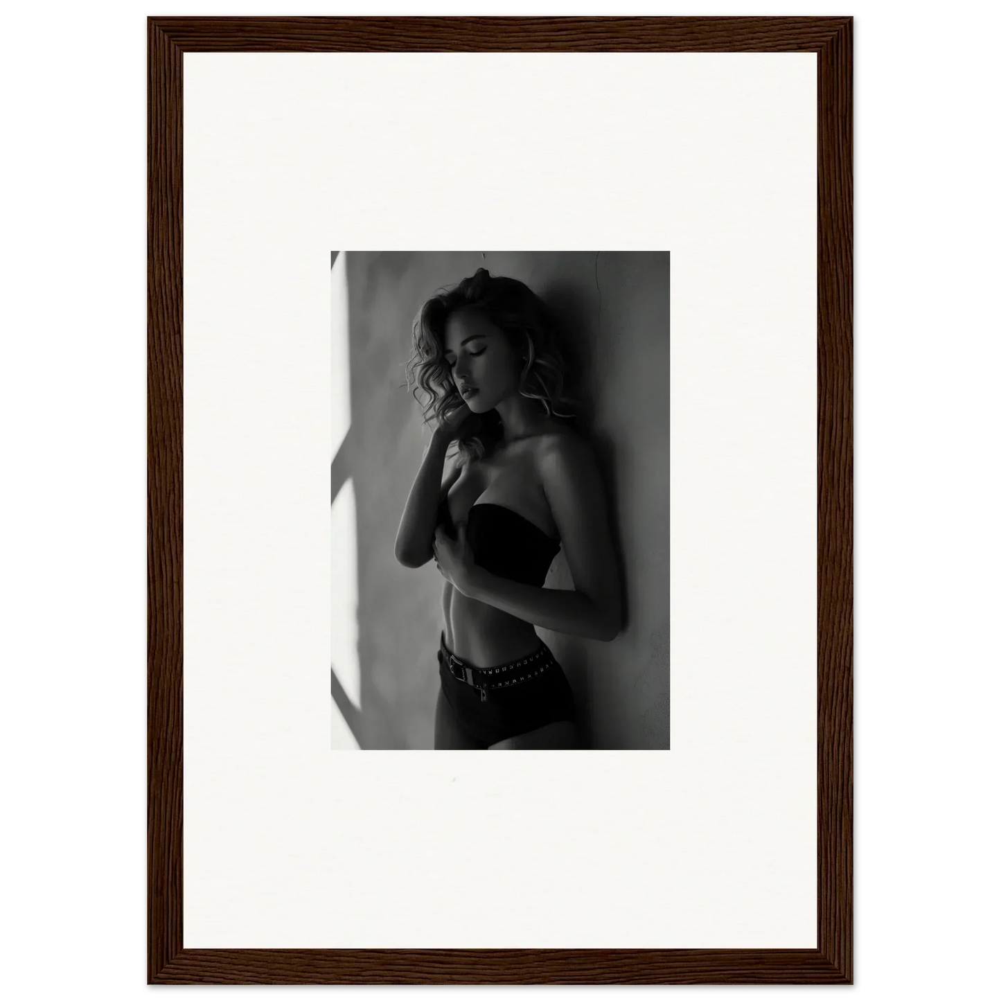 Black and white photograph of a woman in a bra and low-rise pants, posing with one hand near her face.