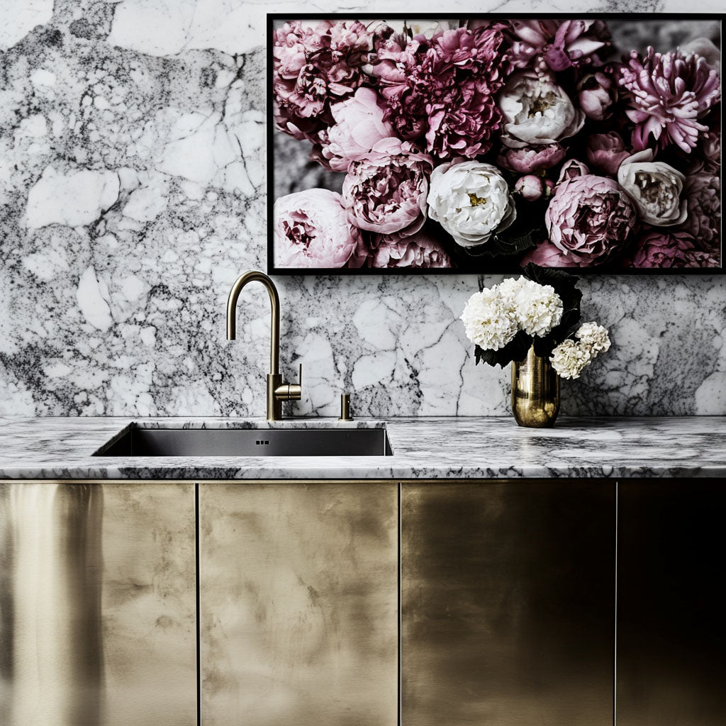 A modern kitchen features a brass sink with a sleek faucet, set against a marbled backsplash. Above it hangs a picture of pink flowers. A vase with white blooms sits on the counter.