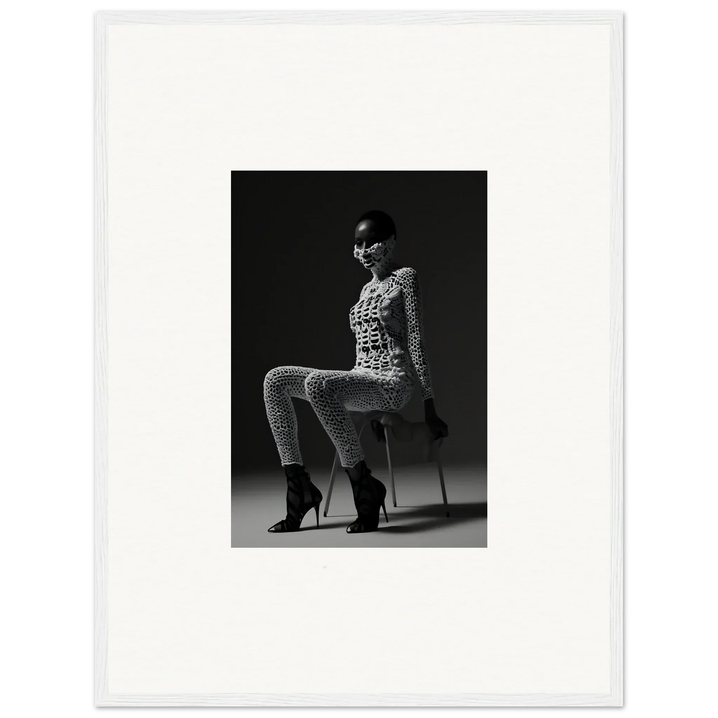 Black and white photograph of a woman in a patterned bodysuit and high heels sitting on an unseen chair.