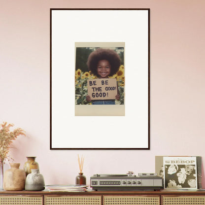 Framed wall art of a person holding a BE THE GOOD sign in sunflower’s gentle whisper