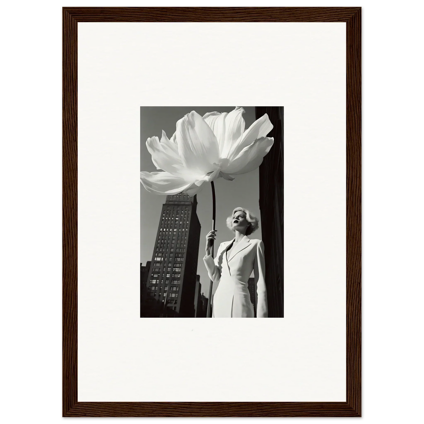 A framed black and white photograph showing a person standing next to an oversized flower against a cityscape backdrop.