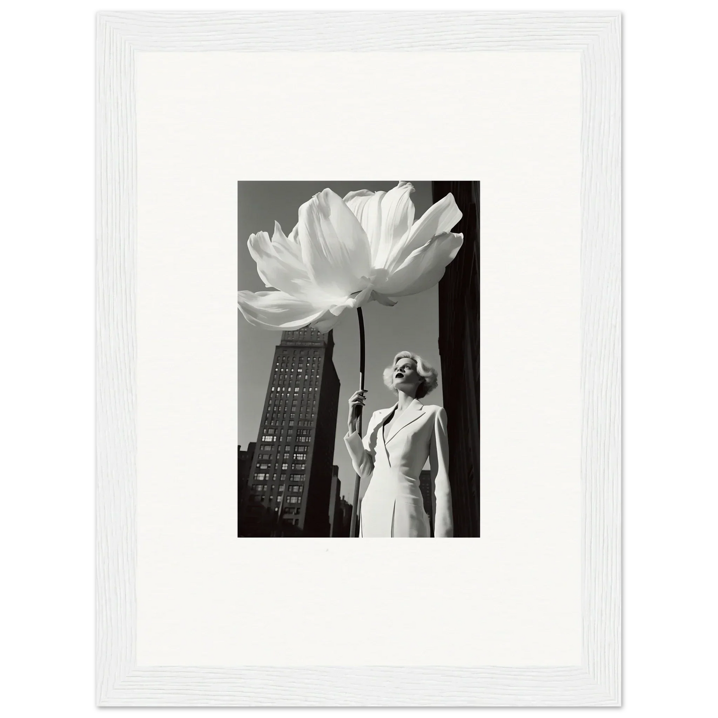 A framed black and white photograph showing a person standing next to an oversized flower against a cityscape backdrop.