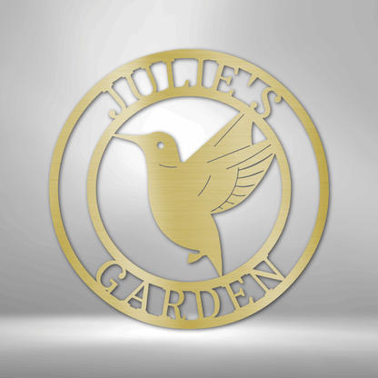 Circular gold-colored metal sign featuring a hummingbird design and the text ’JOHN’S GARDEN’.
