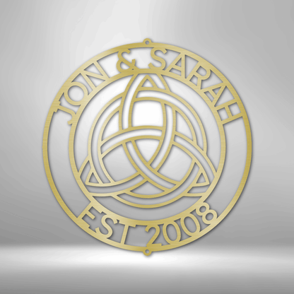 Circular gold-colored emblem featuring a Celtic trinity knot design and text ’JOAN & SARAH EST 2008’’.